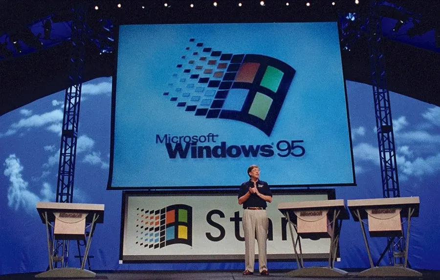 Scena z premiery Windows 95: na wielkim ekranie logo Microsoft Windows 95, przed nim na podeście stoi mężczyzna w ciemnej koszulce, a po bokach niebieska dekoracja z chmurami i stanowiska komputerowe z napisem Microsoft.